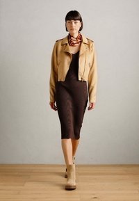 Veste en daim beige courte sur une robe moulante marron à côtes, associée à des bottines beiges et un foulard à motifs autour du cou.