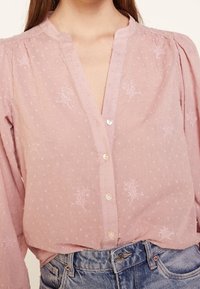 Blusa texturizada rosa con escote en V, botones en el frente y detalles de bordado floral. Presenta mangas abullonadas y una tela suave y ligera.
