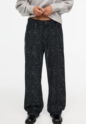 Person wearing loose black leopard-print pants with drawstring waist, a grey sweater lifted to midriff, and black shoes.