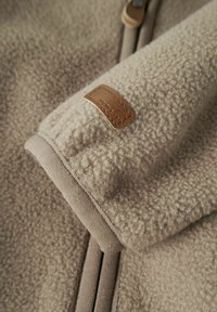 Manica di giacca in pile beige con cucitura visibile e piccola etichetta in pelle marrone del marchio cucita vicino al polsino.