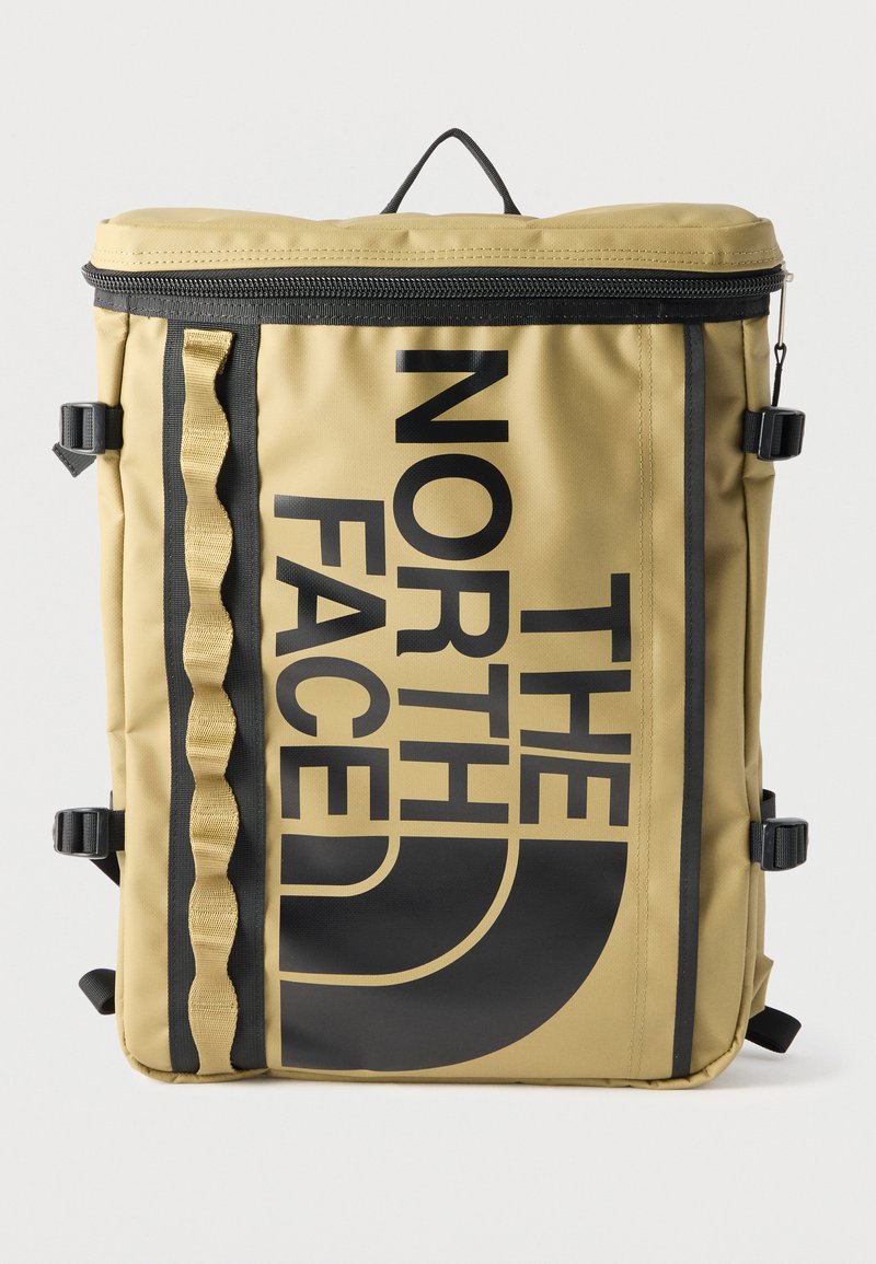 Mochila rectangular de color tostado con cremallera negra, hebillas laterales negras, lazos de cincha y un gran logo vertical negro de "The North Face" en la parte frontal.