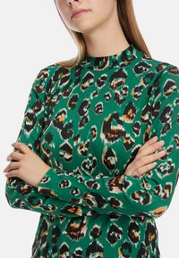 Grüner gerippter Pullover mit einem Leopardenmuster in Schwarz, Orange und Weiß, hohem Kragen und langen Ärmeln.