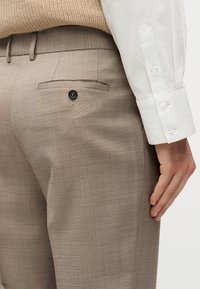 Κοντινό πλάνο μπεζ παντελονιού με τσέπη στο πίσω μέρος με κουμπί, φορεμένο με λευκό πουκάμισο μακρυμάνικο και μπεζ πλεκτό πουλόβερ.