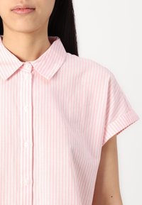 Camisa de manga curta com colarinho em rosa claro com riscas verticais brancas. Características incluem botões e um ajuste descontraído. Tecido macio e texturizado.
