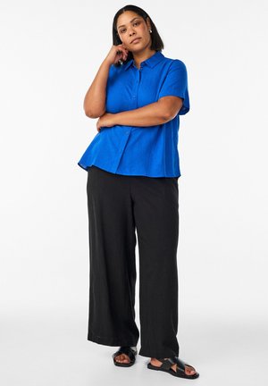 Frau steht mit der Hand am Kinn, trägt ein blaues kurzärmeliges Hemd, schwarze weit geschnittene Hose und schwarze Sandalen vor weißem Hintergrund.