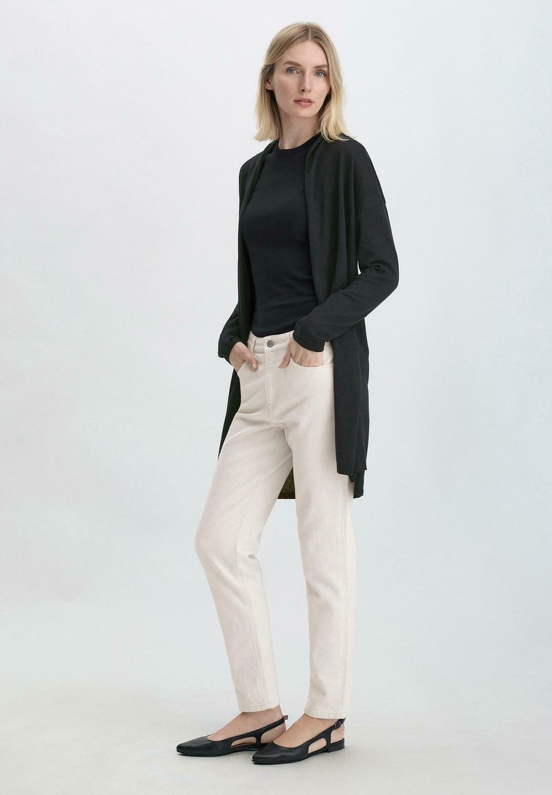 Pitkän mustan neuletakin päällä musta paita, yhdistettynä vaaleanbeigeihin housuihin ja mustiin litteisiin kenkiin. Silkkinen kangas, jossa on minimaalinen sauman yksityiskohta.