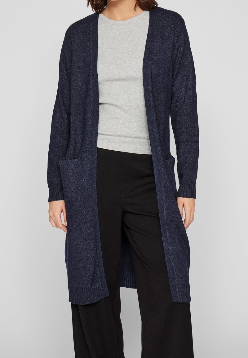 Femme portant un long cardigan bleu marine ouvert sur une chemise gris clair et un pantalon noir à jambes larges, debout devant un fond uni.