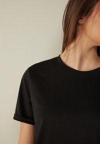 Černé bavlněné tričko s krátkými rukávy a kulatým výstřihem. Hladká textura s ohrnutými rukávy, zdůrazňující jednoduchý design.