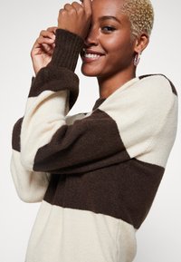 Pull rayé marron et crème avec des poignets côtelés, coupe décontractée et col rond. Texture douce et blocs de couleur marquants.