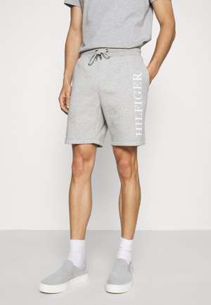 Man in lichtegrijze korte broek met de tekst "HILFIGER" op het linkerbeen, bijpassend grijs T-shirt, witte sokken en grijze instapschoenen.