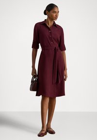 Bordové košeľové šaty s opaskom v páse, vyhrnutými rukávmi a gombíkmi, doplnené malou tmavou kabelkou a zladenými plochými topánkami.