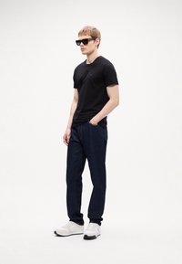 Black short-sleeve t-shirt, dark blue denim pants, white sneakers. Model wearing black sunglasses, standing against a plain background.