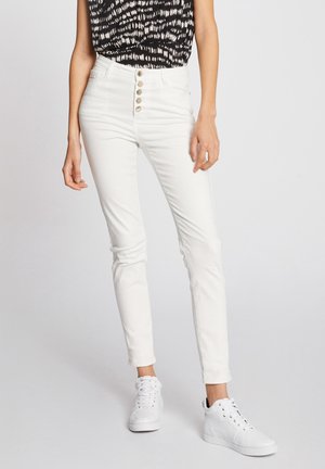 Schmal geschnittene weiße Jeans mit hoher Taille und vier silbernen Knöpfen, kombiniert mit einem schwarz-weiß gemusterten ärmellosen Oberteil und weißen Sneakern.