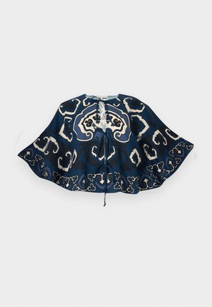 Nő áll, kezét a zsebében tartva, kék mintás, bő felsőt, farmer széles szárú rövidnadrágot és bézs flip-flop papucsot visel egy fehér háttéren.