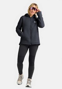 Fleecejack met capuchon in donkergrijs en ritssluiting, gecombineerd met zwarte leggings en lichte wandelschoenen. Voorzien van steekzakken aan de voorkant.