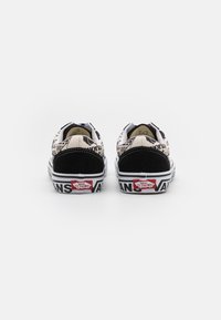 Ett par svarta och vita Vans-sneakers med tidningstryck på hälar och sulor, sedd bakifrån mot en vit bakgrund.