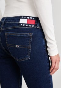 Jeans de denim azul oscuro con una etiqueta "TOMMY JEANS" que presenta acentos en rojo, blanco y azul marino. Los jeans tienen bolsillos traseros y costuras visibles.
