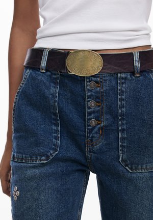 Un haut blanc rentré dans un jean en denim bleu avec une fermeture à boutons et une ceinture en cuir marron foncé avec une boucle en laiton ovale gravée.