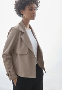 Beżowa kurtka w wzór houndstooth z wyraźnym kołnierzem, klapkami na kieszeniach z przodu i pojedynczym zapięciem na guzik, zestawiona z białą bluzką.