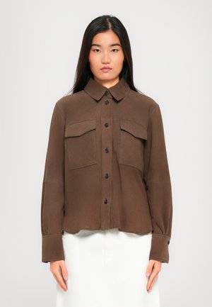 Young woman with long black hair wears a brown buttoned shirt with flap pockets and white skirt, standing against a plain light background.