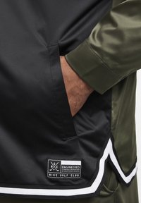 Czarna i oliwkowa kurtka z boczną kieszenią, gładką teksturą oraz naszywką z etykietami "Nike Golf Club" i "Engineered", z białymi akcentami.