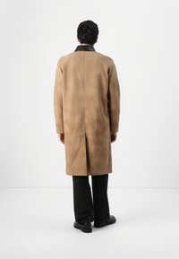 Un long manteau beige en daim, doté d'un col en cuir noir et d'une coupe droite, porté sur un pantalon noir et des chaussures.