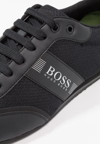 Black athletic shoe featuring a mesh upper, smooth leather accents, and a prominent white "BOSS" logo on a stripe. Laces are dark woven fabric.