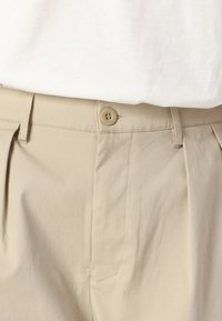 Pantalons beige à coupe droite, plis à la taille et fermeture à un seul bouton. Fabriqués en tissu lisse et léger avec un minimum de coutures.