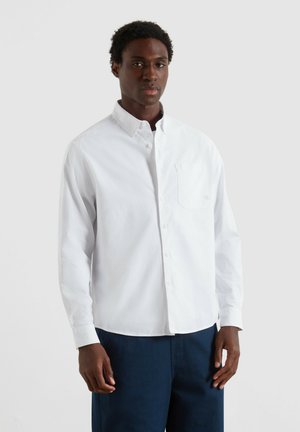 Uomo con camicia bianca a maniche lunghe e pantaloni blu scuro, in piedi davanti a uno sfondo grigio chiaro uniforme.