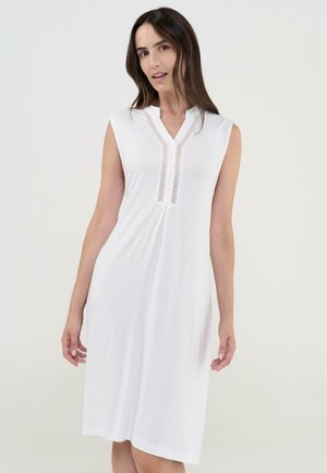 Mujer con vestido blanco sin mangas hasta la rodilla, con escote en V abotonado y adorno vertical decorativo, de pie contra un fondo liso.