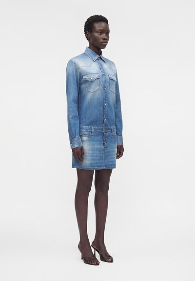 Γυναίκα που φορά ξεθωριασμένο φόρεμα πουκάμισο από τζιν με τσεπάκια στο στήθος με κουμπιά και ψηλοτάκουνα παπούτσια ανοιχτά μπροστά, στέκεται σε λευκό φόντο.