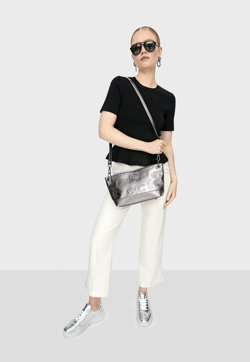 Borsa a tracolla argento con logo in rilievo, abbinata a un top nero a maniche corte, pantaloni bianchi a gamba larga e sneakers argento. Texture liscia, design moderno.