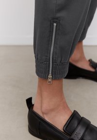 Alara jalkaan pukeutuneet tummanharmaat housut, joissa on hopeinen vetoketju lähellä elastista nilkkaosaa, yhdistettynä mustiin liukusapatoihin vaalealla lattialla.