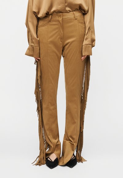 Pantalon marron à franges avec des fermetures éclair latérales et une texture lisse, associé à des ballerines noires à bouts pointus. Le pantalon a une coupe droite.