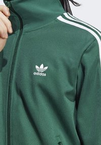 Grön zip-up-jacka i texturerad tyg, med vita tre-stripes detaljer på ärmarna och en vit Adidas-logga på bröstet.