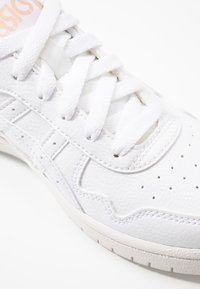 Weiße Sneaker mit Obermaterial aus strukturiertem Leder, kontrastierender kremfarbener Mittelsohle und flachen weißen Schnürsenkeln. Zeigt Perforationen und ein stilisiertes Seitenmuster.