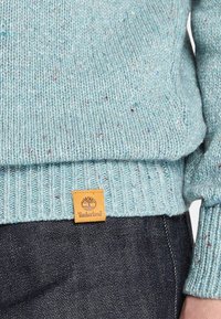 Jasnoniebieski sweter z dzianiny o fakturowanym wzorze, z prążkowanym dołem i brązową naszywką z logo Timberland w pobliżu dolnej krawędzi.