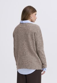 Rjavi puhasti pulover z ohlapnim prileganjem, okroglim izrezom, dolgimi rokavi in svetlo modro srajco, ki kiti izpod puloverja.