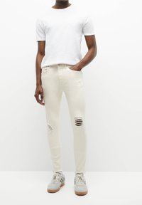 Jeans ajustados de color claro con roturas, material de algodón, que presentan múltiples áreas desgastadas. Combinados con una camiseta blanca sencilla y zapatillas grises.