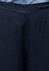 Κοντινό πλάνο σκούρου μπλε υφασμάτινου παντελονιού που φοριέται με ανοιχτό μπλε πουκάμισο με κουμπιά, μαζεμένο στη μέση.