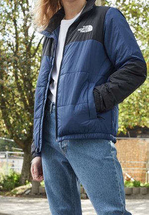 Person wearing blue and black The North Face jacket and blue jeans, standing outdoors near trees on a sunny day.