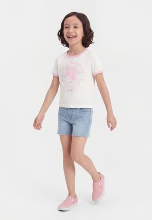 Χαμογελαστό παιδί με κοντά σγουρά μαλλιά που περπατά, φοράει λευκό T-shirt με ροζ λεπτομέρειες και στάμπα, ανοιχτό μπλε τζιν σορτς και ροζ παπούτσια χωρίς κορδόνια.