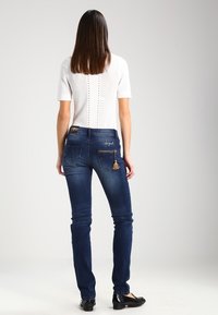 Jean skinny en denim bleu foncé, présentant deux poches arrière avec des zips et un détail de frange, associé à un top blanc à manches courtes.