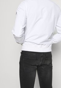 Chaqueta bomber blanca con puños y dobladillo acanalados, combinada con jeans negros que tienen bolsillos traseros. Tejido suave, diseño casual.