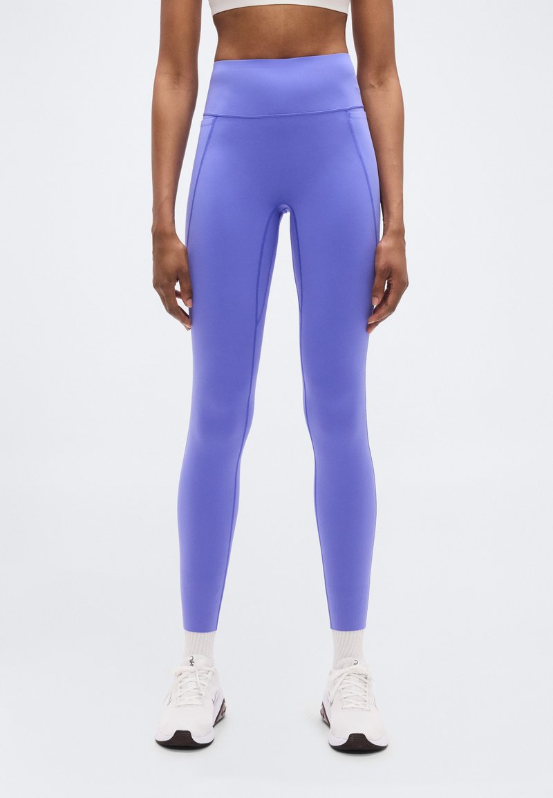 Κολάν με ψηλή μέση, εφαρμοστά, σε μονοχρωμία μωβ με λείο, ελαστικό ύφασμα. Συνδυασμένο με λευκά αθλητικά παπούτσια και κανονικές κάλτσες.