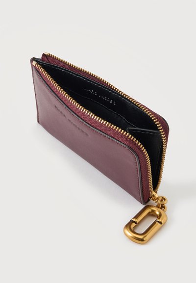 Petite pochette en cuir bordeaux avec une fermeture éclair dorée et un grand porte-clés doré fixé, montrée partiellement ouverte sur une surface blanche.