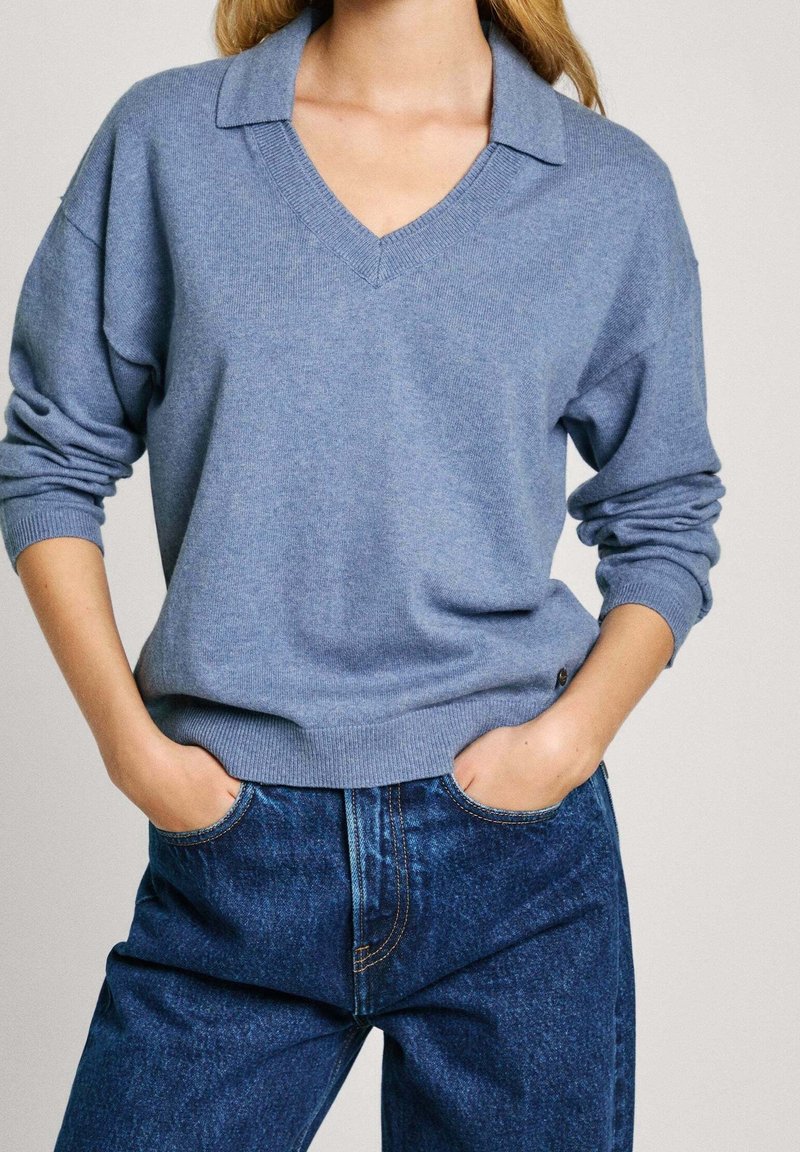 Femme portant un pull léger bleu clair à col en V avec manches longues et un jean en denim bleu, les mains glissées dans les poches avant.