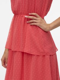 Gros plan sur une personne portant une robe texturée corail rose avec un détail de taille à basque, une main reposant sur la hanche et l'autre détendue.