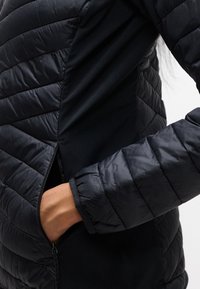 Χέρι στην τσέπη μαύρου καπιτονέ μπουφάν με το φερμουάρ μερικώς ανοιχτό, αποκαλύπτοντας λείο ύφασμα στο εσωτερικό.