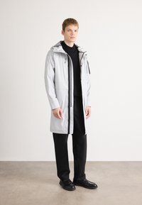 Jachetă impermeabilă gri deschis, cu glugă, fermoare negre și buzunare. Purificată peste un pulover negru și pantaloni negri, având un design drept.